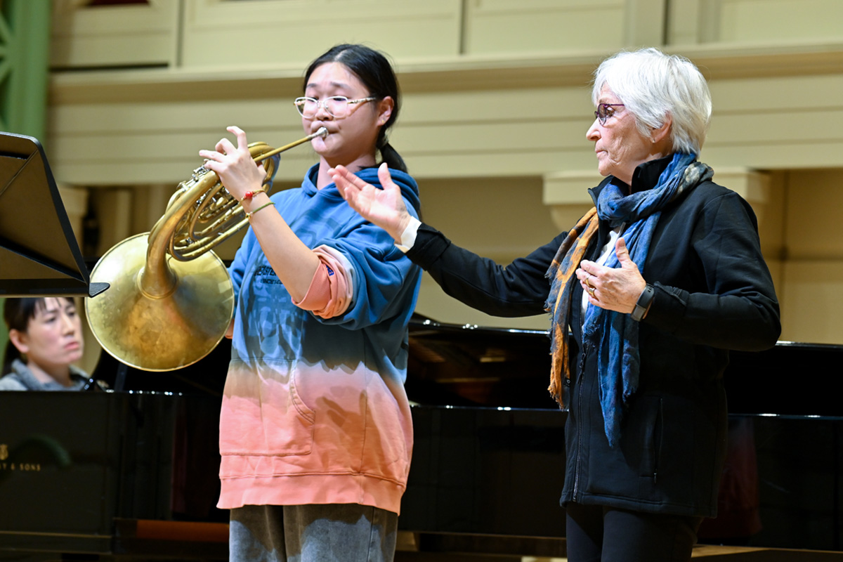 A female professor leading a female horn player in a music performance on stage.